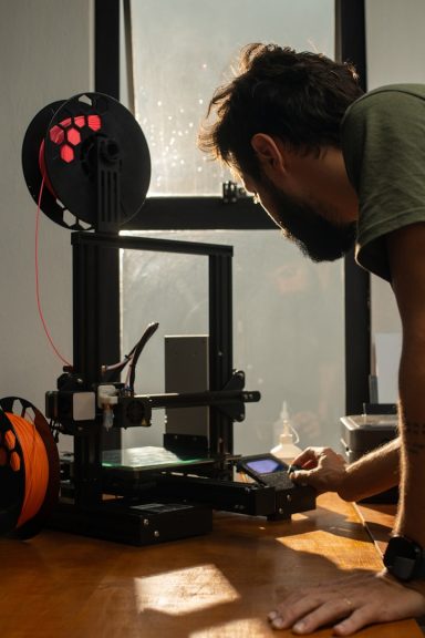 Mann arbeitet an einem 3D-Drucker am Tisch, mit Sonnenlicht im Hintergrund.
